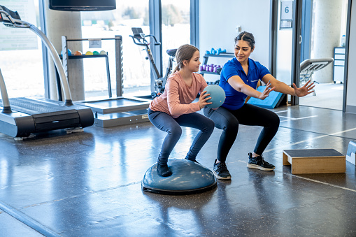 A young girl works with her physiotherapist in a gym during an appointment.  She is dressed comfortably and is balancing on a half ball as she squats down with a ball in her hands.  The therapist is spotting her as she repeats the exercise.