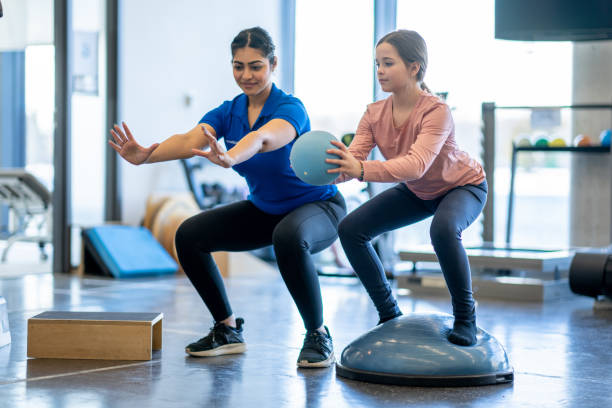 Children's Physiotherapy A young girl works with her physiotherapist in a gym during an appointment.  She is dressed comfortably and is balancing on a half ball as she follows the therapist instructions and does a squat with a ball in her hands. sports medicine stock pictures, royalty-free photos & images