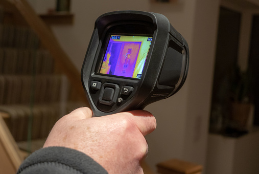 A male surveyor using a handheld infrared thermal camera, to inspect the interior of a domestic home, looking for heat anomalies. The camera viewfinder shows a coloured thermal image with varying temperature ranges.