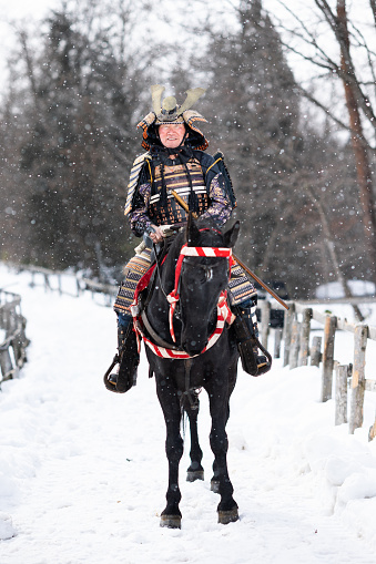 A retired senior man in samurai armor riding his trusty horse along a snowy farm path.