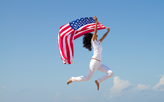 4th July concept: young active woman holding american flag and jumping on beach