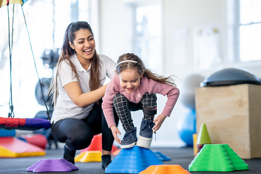 A female physical therapist helps a little girl with a limb difference through a therapy course.  The little girl is balancing on a block as she navigates the colorful course and works on balance with her brace on.