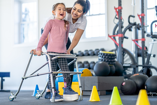 A little girl with a limb difference and a leg brace works with her physical therapist as she learns to use a walker.  The female therapist is bending down to aide the little girl as she directs her through a course.