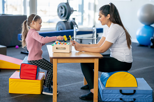 A little girl with a limb difference and a leg brace, sits at a table with her therapist as they work on her speech.  The therapist is using colorful blocks and activities to make the therapy fun.