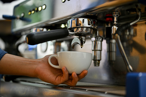 Close-up image of a professional male barista preparing customer's order, making a cup of cappuccino with coffee espresso machine.