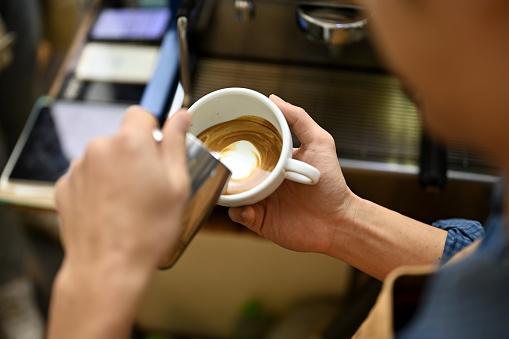 Close-up image of talented Asian male barista pouring steamed milk into a cup, making latte art, working in cafe.