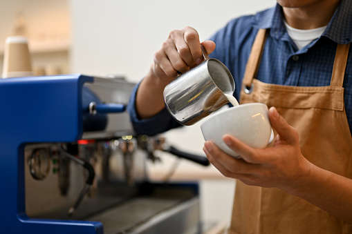 Cropped shot of Asian male barista in apron pouring steamed milk into a cup, making hot latte or cappuccino.