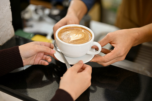 Close-up image of a male barista handing a cup of latte to a customer in the coffee shop.