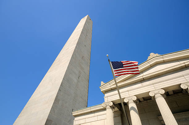 Bunker Hill Monument, Boston, Massachusetts. Low angle view of obelisk, building facade and American Flag against clear blue sky.