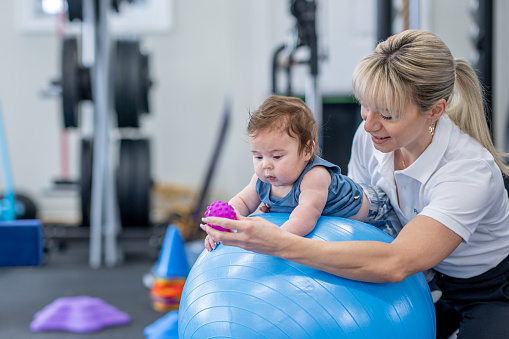 A female physical therapist works with an infant during a therapy session.  She has the baby boy laying on a large ball on his tummy as she helps him work on his core and neck strength.