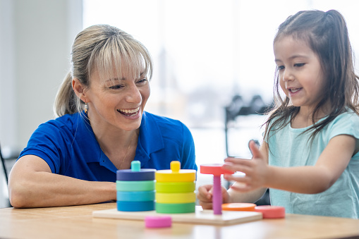 A female therapist sits with a little girl at a table as they work on her motor skills.  The therapist is dressed professionally and is using colorful toys to help her build strength in her hands and coordination.