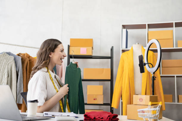 A beautiful blogger shows off her clothes in front of a smartphone camera while recording videos, vlogs and live streams at her store, online clothing sales concept on social media.