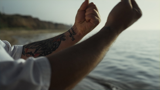 Tattooed man hands doing warm-up at sunset outdoors close up. Unknown sportsman making sporty exercises standing on beach. Strong muscular arms rotating on sunlight. Workout on summer nature.