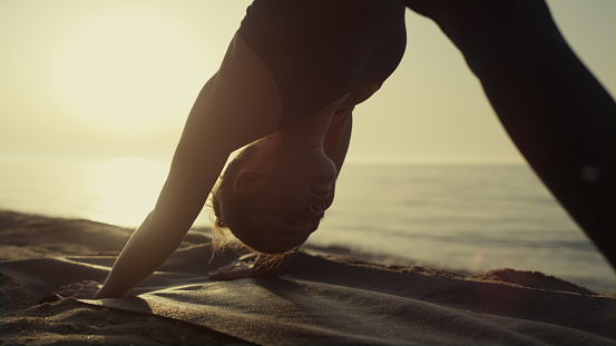 Yogi girl practicing asana standing pose down face dog on sandy beach close up. Attractive young woman stretching at sunset summer time. Focused sportswoman training flexibility on mat outdoors.
