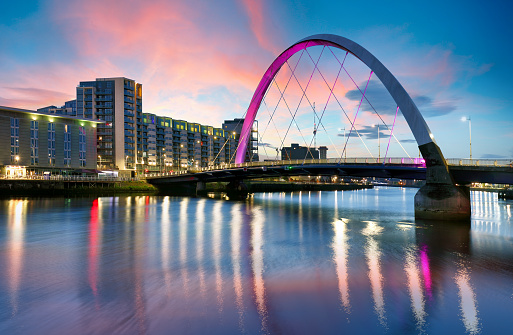 Beautiful Sunset Clyde Arc Bridge across river in Glasgow, Scotland, UK. It is nice weather with reflection on water, blue sky, lights from buildings in downtown, skyline, attractions.