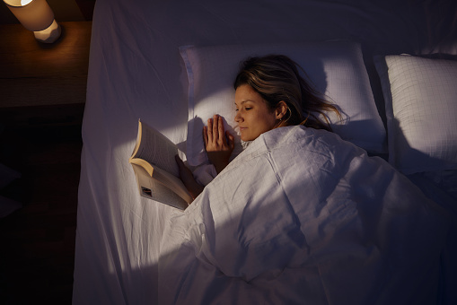 High angle view of young woman reading a book while relaxing in a bed during the night.