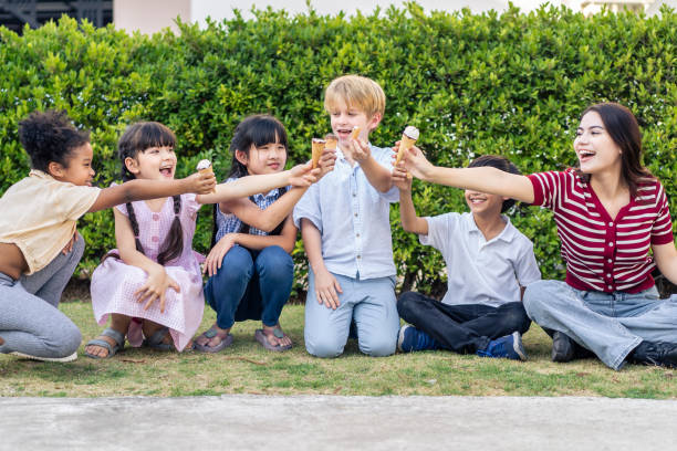 Group of young children eating an ice cream while having a picnic outdoors with teacher together in garden. Adorable boys and girls kid feel happy and relax, enjoy fun activity at elementary school. grupo de niños pequeños comiendo un helado mientras hacen un picnic al aire libre con el maestro juntos en el jardín. adorables niños y niñas niños se sienten felices y se relajan, disfrutan de actividades divertidas en la escuela primaria.