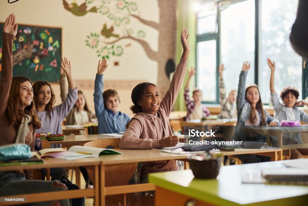 Happy elementary students raising their hands on a class at school. Large group of happy elementary students know the answer on a class in the classroom. Focus is on black girl. Child Stock Photo