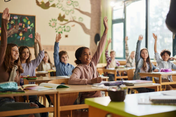 Happy elementary students raising their hands on a class at school. stock photo