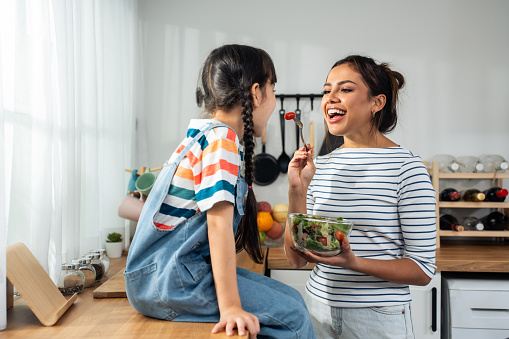 Asian mother teach and motivate young girl child eat green vegetable. Adorable little kid child feel happy and relax, enjoy eat salad and healthy foods for health care and wellness in kitchen at home.