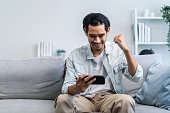 Asian young handsome man gamer play mobile game on smartphone at home. Attractive casual guy feel happy and relax, sit on sofa having fun touching screen on phone to play video enjoy victory in house.