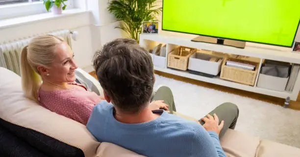 Couple watching green screen TV while sitting on sofa in living room.