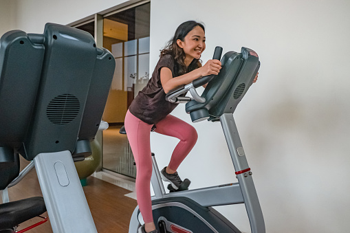 Side shot of thin Southeast Asian woman with a protective face mask, doing cardio exercise above elliptical cross trainer machine at empty gym