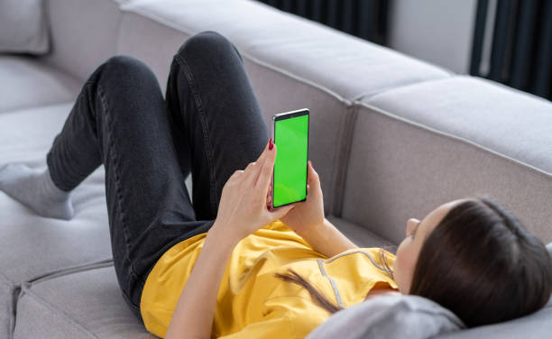 istock Feminine Hand Scrolling Feed on Smartphone with Green Screen Mock Up Display. Female is Relaxing on Sofa at Home, Watching Videos and Reading Social Media Posts on Mobile Device. 1468144452