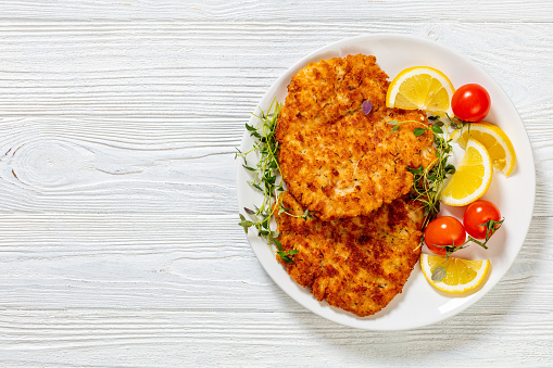 fried juicy chicken breast cutlets under crusting of panko breadcrumbs on white plate with lemon, fresh thyme and ripe tomatoes on white wood table, flat lay, copy space