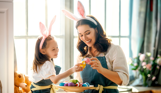 Happy mother and her daughter wearing aprons holding painted colorful eggs while decorating them with food dyes in cozy kitchen at home. Easter craft activities for families.