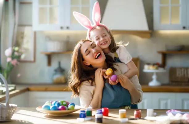 Happy mother and her daughter wearing aprons holding painted colorful eggs while decorating them with food dyes in cozy kitchen at home. Easter craft activities for families.