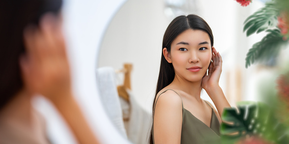 Portrait of attractive young woman looking at mirror. Beautiful girl enjoying her reflection, self-care concept.