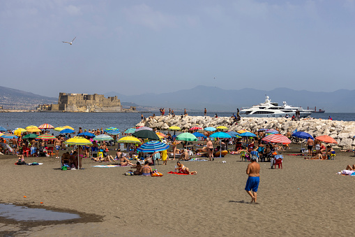 Naples, Gulf of Naples, Italy - June 27, 2021: People relaxing on the Mappatella Beach by the Tyrrhenian Sea next to the boulevard Chiaia, colorful umbrellas, Castel dell'Ovo in the background