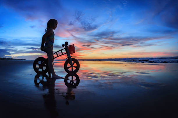 Black silhouette of little child - run bike rider stand on wet sand beach, look at sea surf and colourful sunset sky. Family travel lifestyle background, outdoor fun activity on Bali summer vacation.
