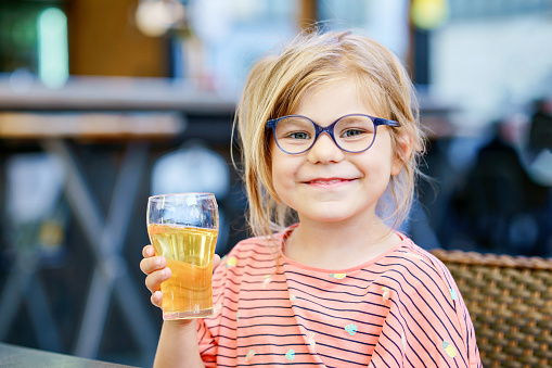 Adorable little girl having breakfast and drinking juice at resort restaurant. Happy preschool child with glasses holding glass with apple juice.