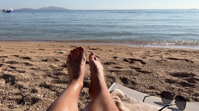 Young beautiful sexy tanned girl in bikini lying on the beach by the sea. View of the sea through the female legs, feet