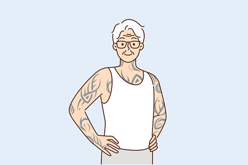 Optimistic elderly man with tattoos on arms and body stands with hands on belt proud good health in old age. Gray-haired elderly human with tattoos stuffed in youth looks at screen posing in t-shirt