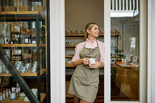 Front view of cheerful woman wearing apron over casual attire, standing in doorway of specialty store, holding mug and looking away.
