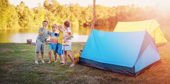 Group of four children standing near colourful tents in nature. Concept of the family vacation, recreation and scouts camping.