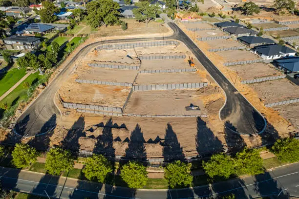 Aerial view new rural housing development earthworks, retaining walls, access roads with display homes to right of frame. Late afternoon shadows from a row of tapered conifer trees seem to point to the new development. A tree-lined boulevard in the foreground provides a thoroughfare for the hundreds of new homes in the area. Mount Barker, South Australia