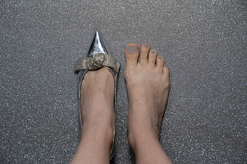 female feet with silver fashion shoe, woman wears heels on one leg, second leg barefoot with pedicure, concept of trying shoes in the store