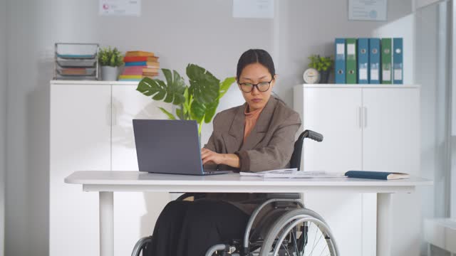 Young pretty disabled woman in wheelchair working on laptop computer at office.