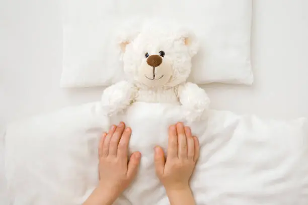 Photo of Little child hands putting white fluffy teddy bear to sleep on pillow and sheet under blanket in baby crib. Closeup. Point of view shot. Kids best friend. Top down view.