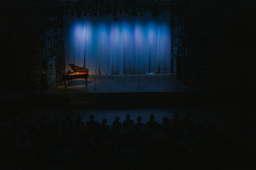 music stage theater with grand piano and white backdrop illuminated with stage light and audience in silhouette