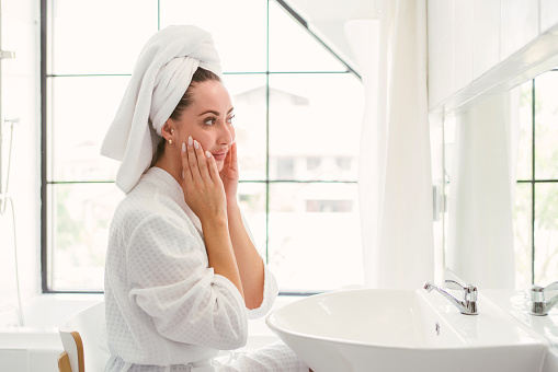 Gorgeous confident adult woman wearing a towel on her head sitting and checking her face in front of the mirror in the home bathroom. A pretty attractive woman beauty lifestyle and cosmetic concept.