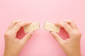 istock Young adult woman hands holding broken wafer pieces on light pink table background. Pastel color. Sweet snack. Two pieces. Closeup. Point of view shot. Top down view. 1468189774
