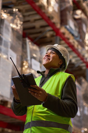Vertical portrait of black woman wearing hardhat in storage warehouse and smiling happily