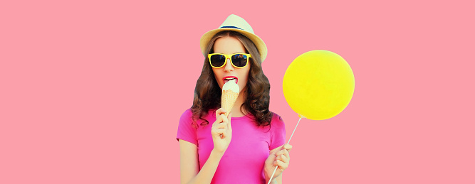 Portrait of young woman eating ice cream holding yellow balloon wearing summer straw hat on pink background