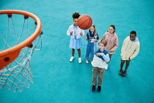 Above angle of intercultural schoolchildren playing basketball on playground together while one of them throwing ball into basket