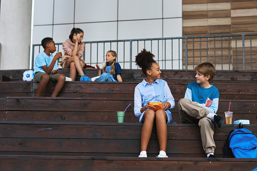 Two happy intercultural schoolmates sitting on wooden staircase at lunch break, having chat and eating sandwiches against group of kids